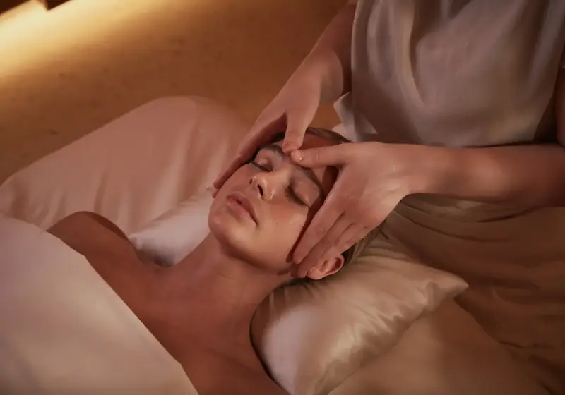 A person lies on a massage table with eyes closed while a therapist gently massages their forehead and temples using both hands in a softly lit spa setting.