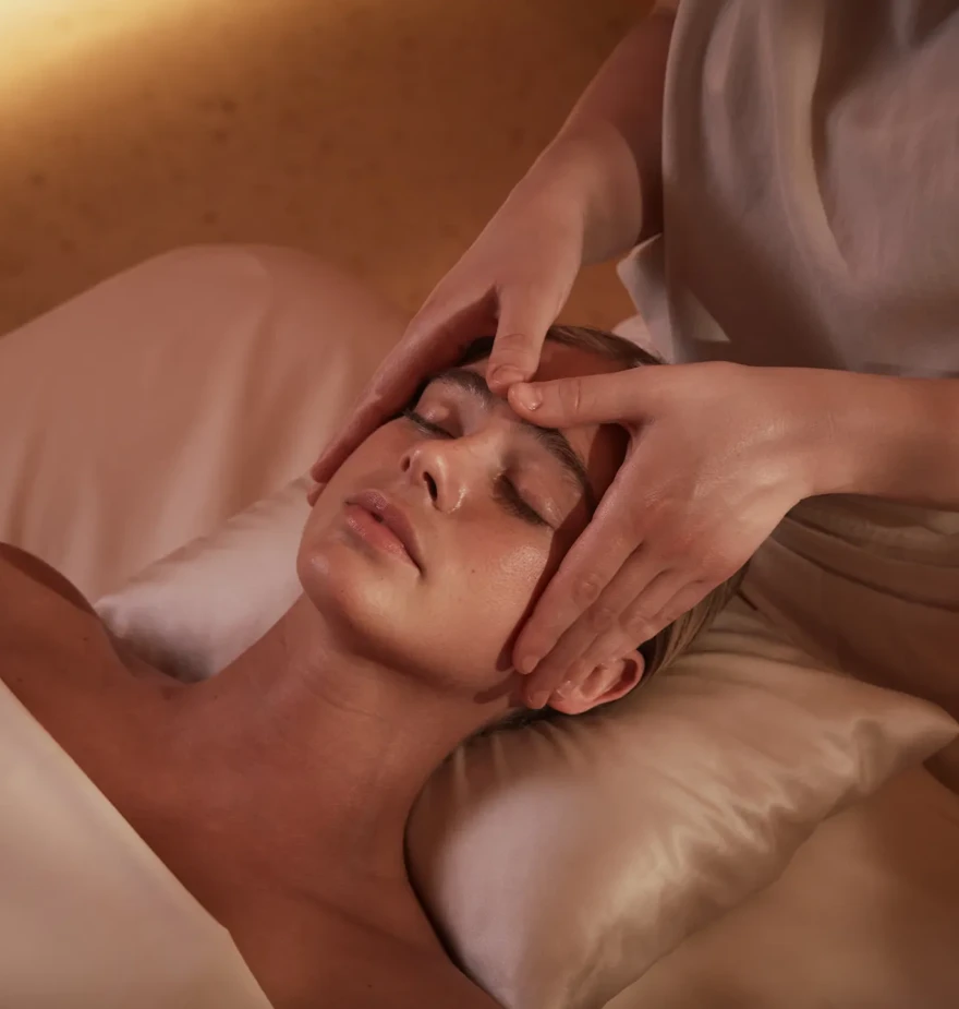 A person lies on a massage table with eyes closed while a therapist gently massages their forehead and temples using both hands in a softly lit spa setting.