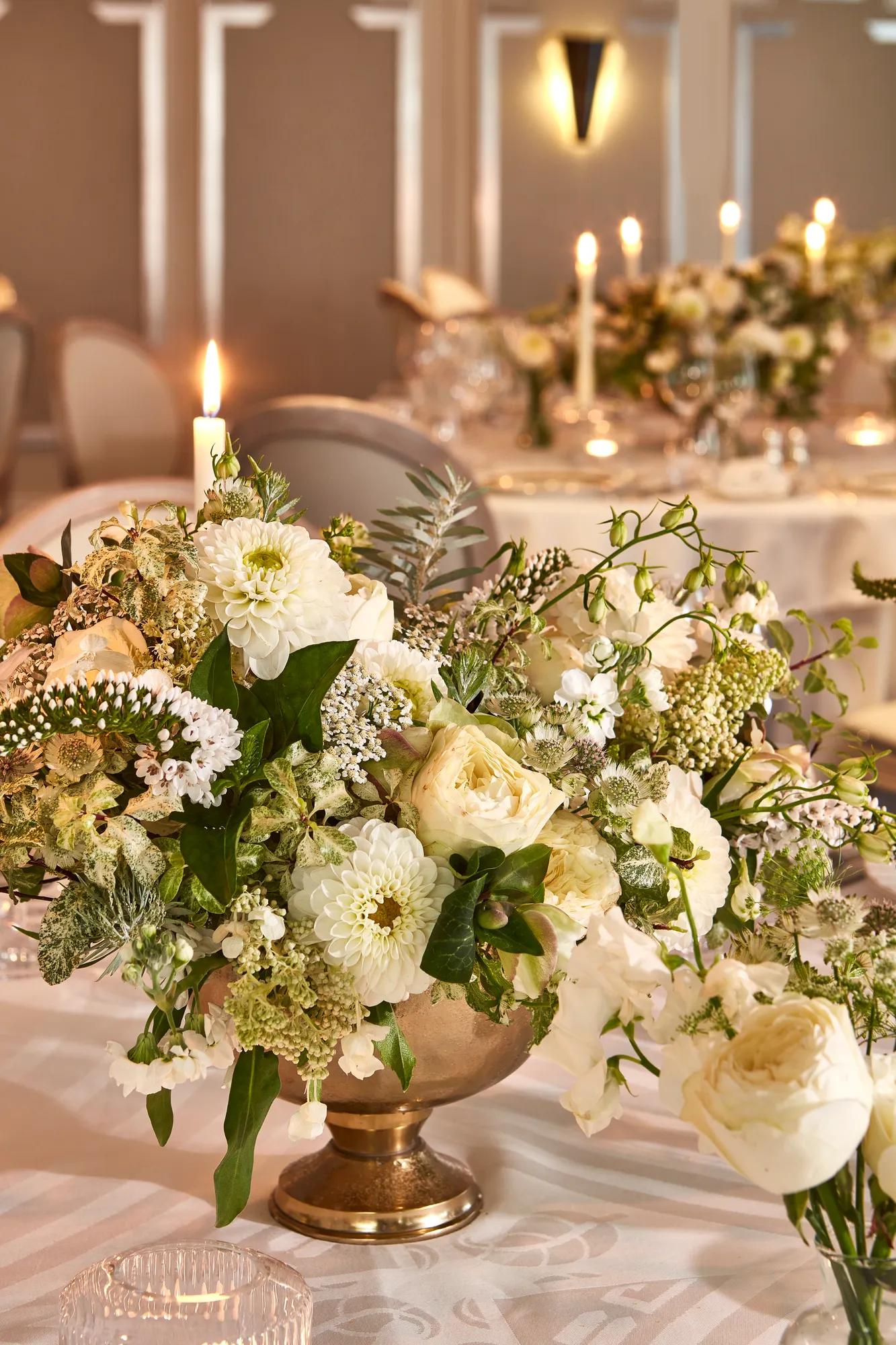 Elegant white floral centrepiece in gold vase on a candlelit dining table, with soft lighting and blurred tables set for a formal event in background.