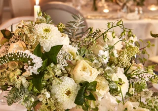 Elegant white floral centrepiece in gold vase on a candlelit dining table, with soft lighting and blurred tables set for a formal event in background.