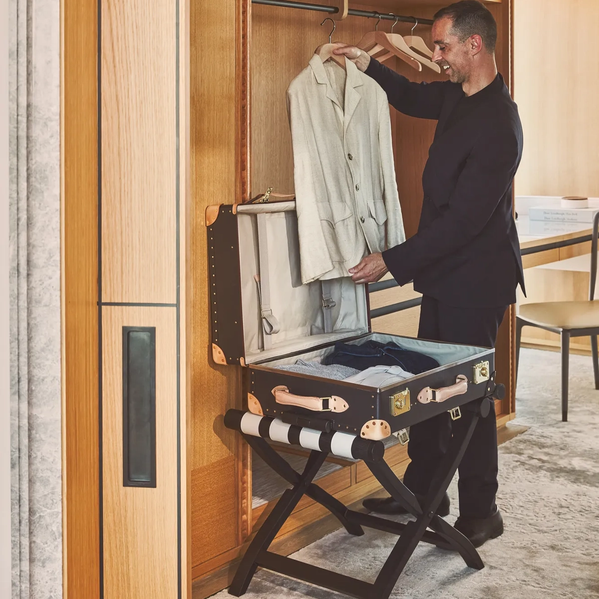 Hotel staff member smiling while unpacking a beige jacket from a luxury trunk in a suite wardrobe.