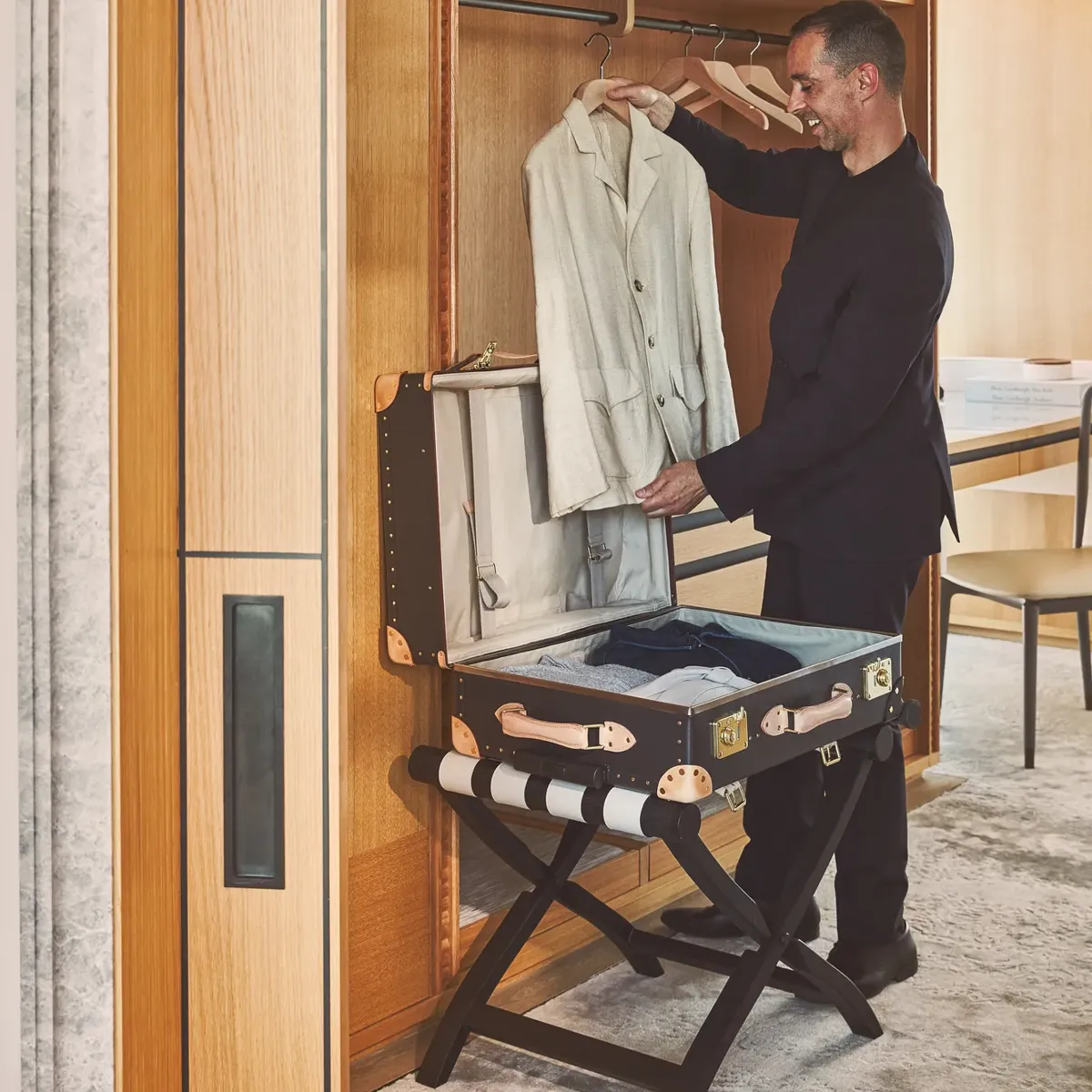 Hotel staff member smiling while unpacking a beige jacket from a luxury trunk in a suite wardrobe.