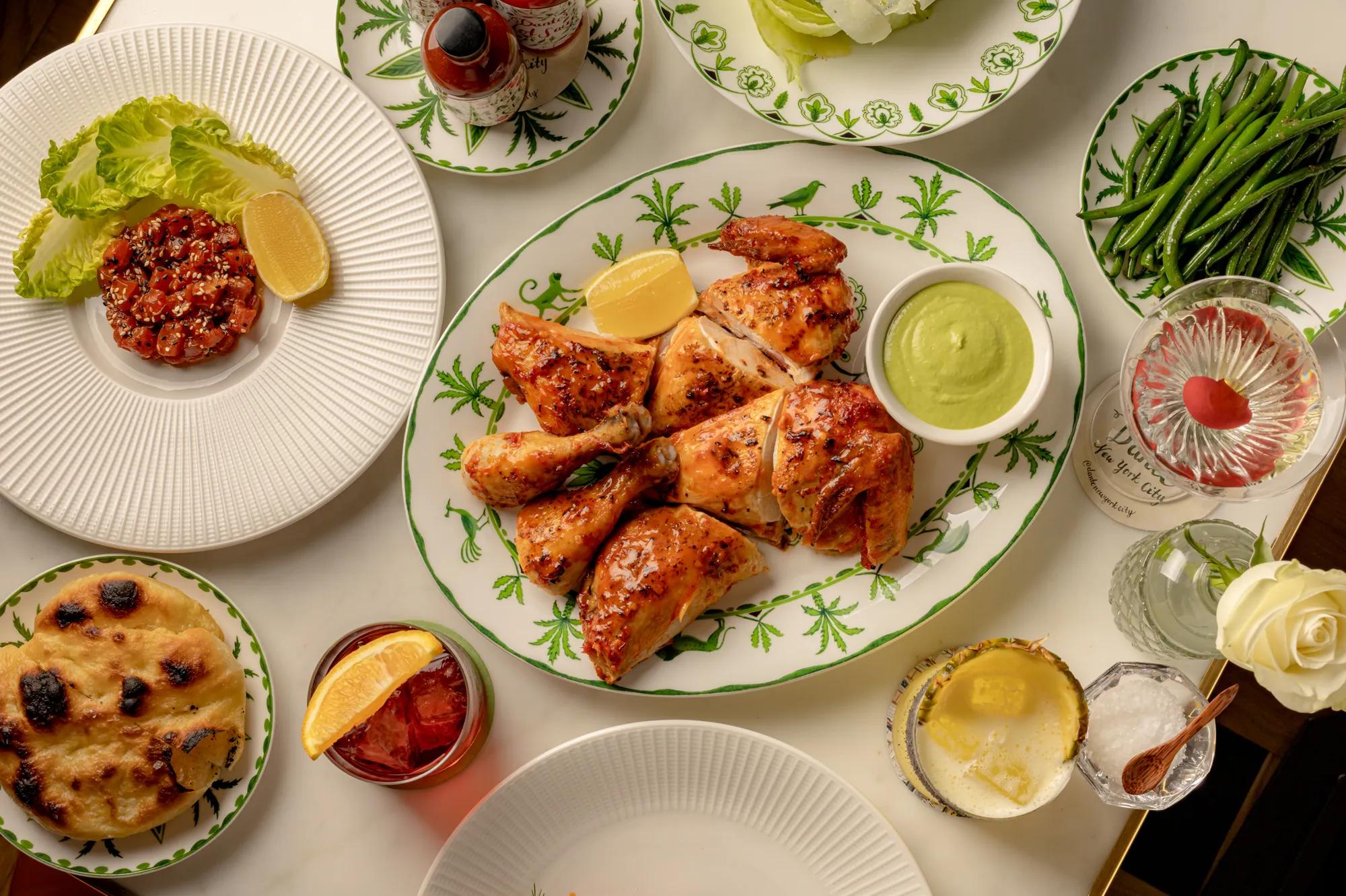 Grilled chicken pieces served on a patterned platter with green sauce, surrounded by salads, green beans, flatbread and cocktails on a dining table