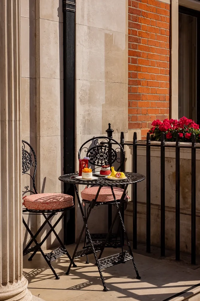 Small outdoor bistro table with pastries and drinks on a sunny terrace, beside two ornate black chairs and flower-filled railing.