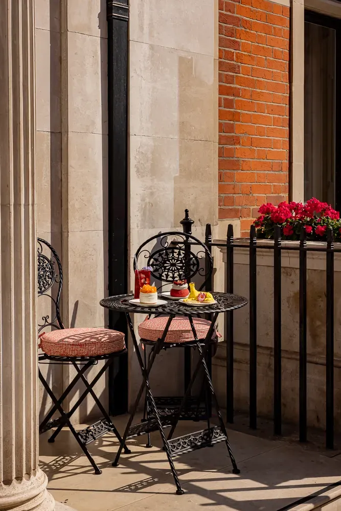 Small outdoor bistro table with pastries and drinks on a sunny terrace, beside two ornate black chairs and flower-filled railing.