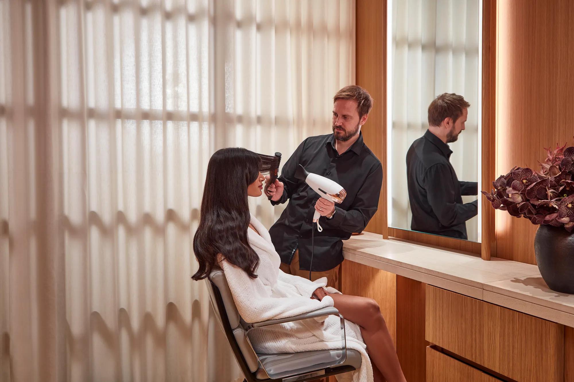 Stylist blow-drying a guest’s hair at a salon vanity, with mirror, wood panelling and soft natural light.