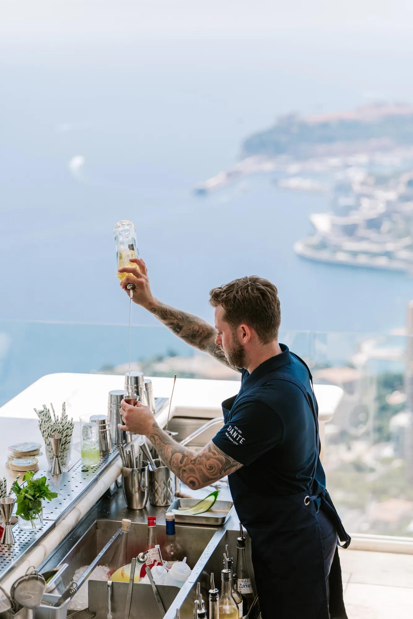 Bartender pouring a drink at an outdoor bar with panoramic coastal views and marina in the background.