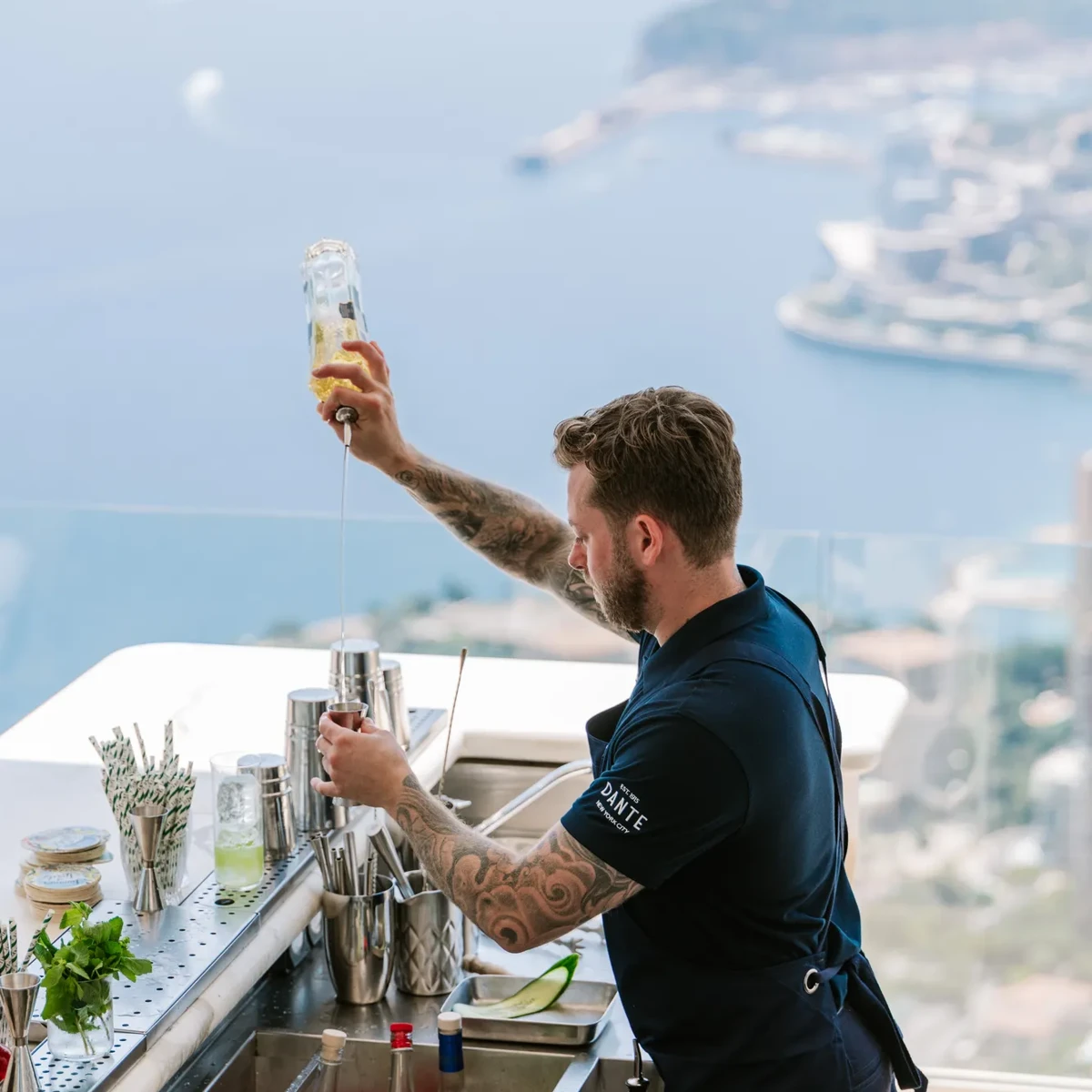 Bartender pouring a drink at an outdoor bar with panoramic coastal views and marina in the background.