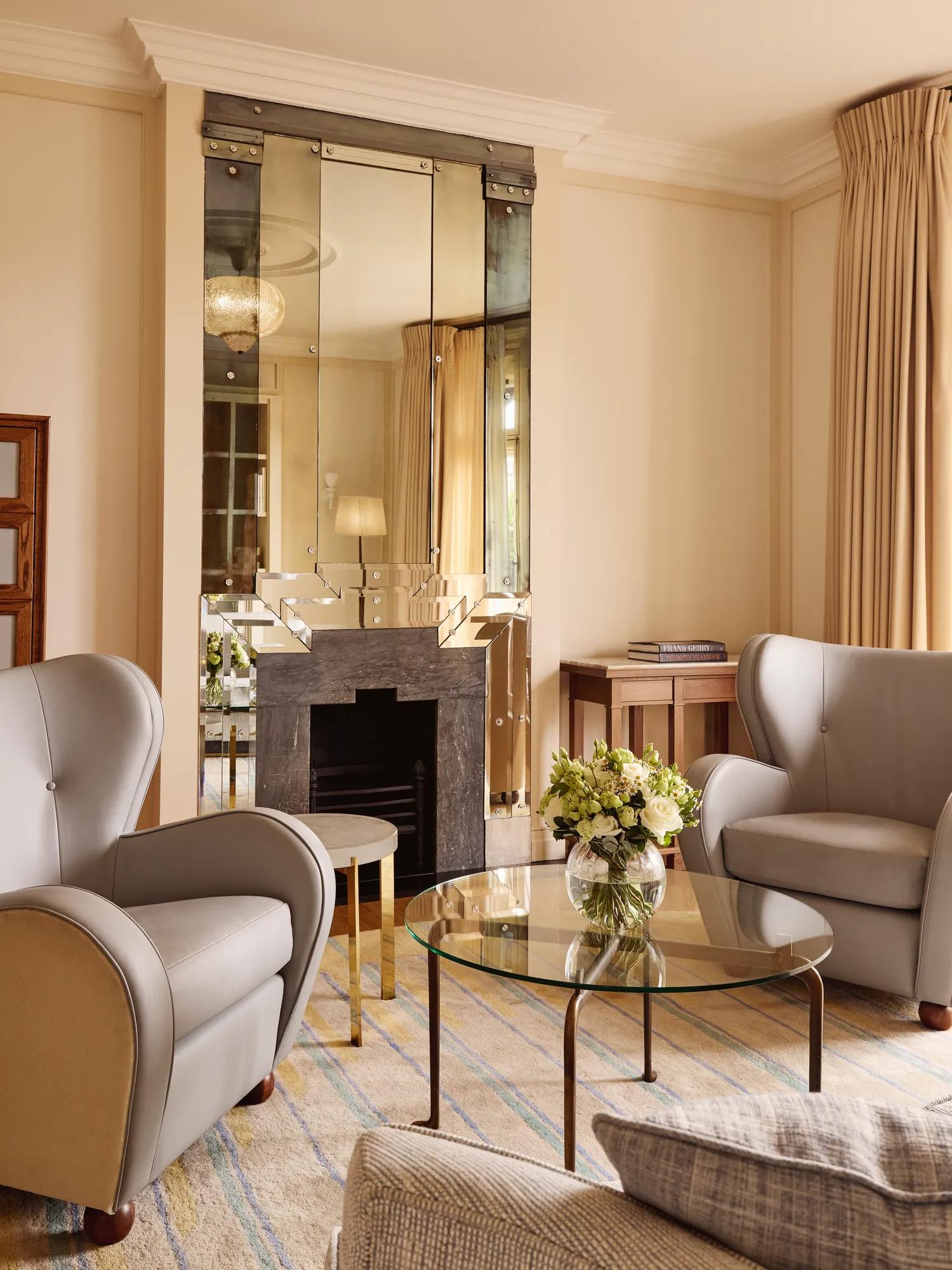 Refined sitting area with armchairs, glass coffee table, floral arrangement, and decorative fireplace beneath a mirrored feature wall.