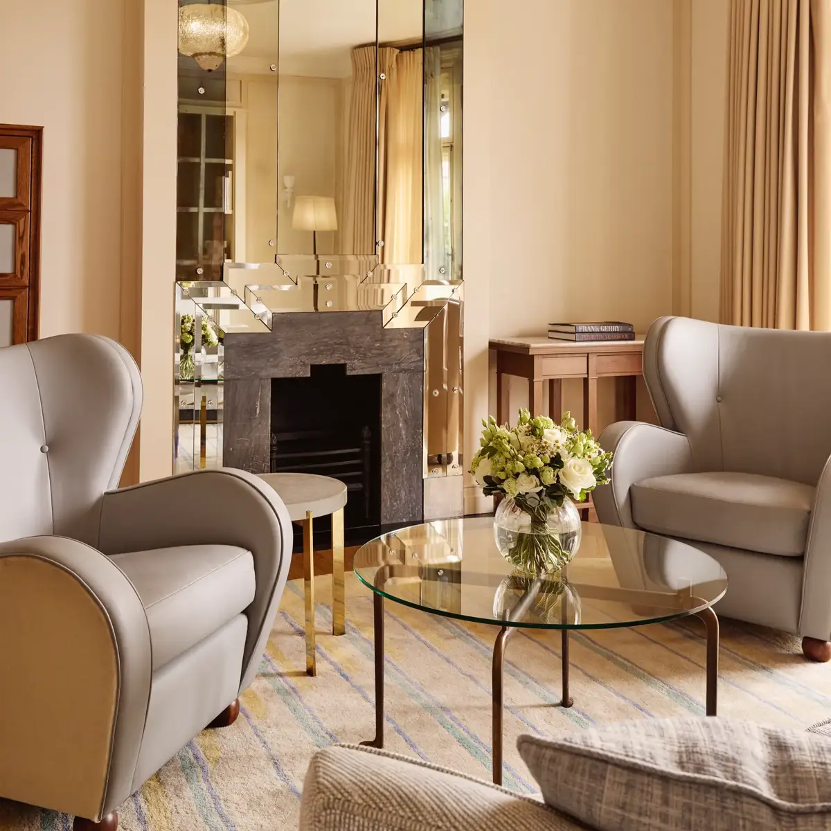 Refined sitting area with armchairs, glass coffee table, floral arrangement, and decorative fireplace beneath a mirrored feature wall.
