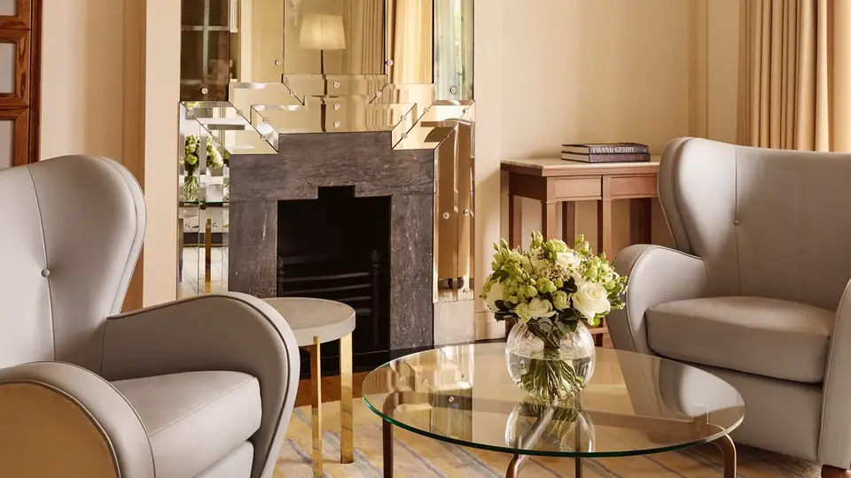 Refined sitting area with armchairs, glass coffee table, floral arrangement, and decorative fireplace beneath a mirrored feature wall.
