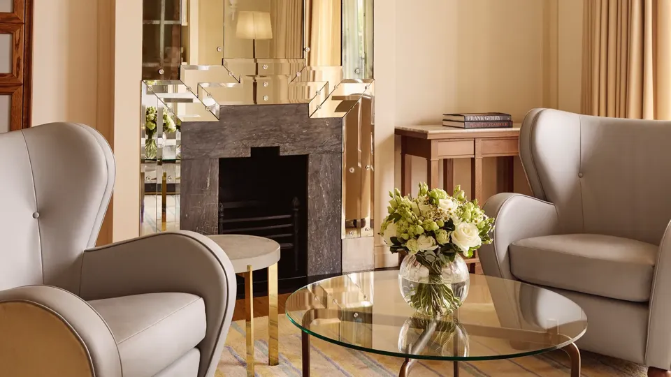 Refined sitting area with armchairs, glass coffee table, floral arrangement, and decorative fireplace beneath a mirrored feature wall.