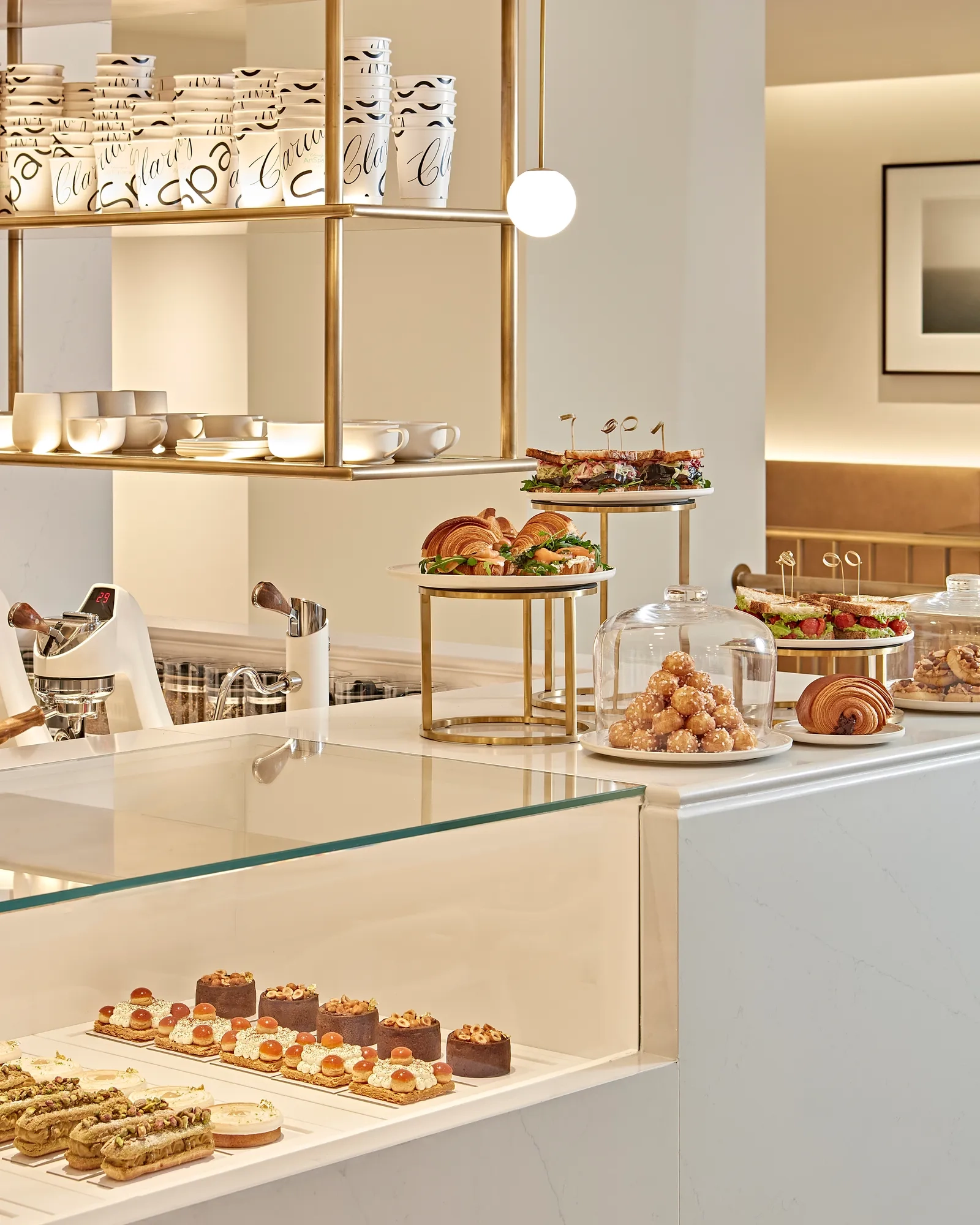Bright and elegant patisserie counter displaying an assortment of pastries, cakes, and sandwiches on glass and tiered stands. The white and gold interior features modern shelving, ceramic cups, and soft lighting that highlights the refined presentation.