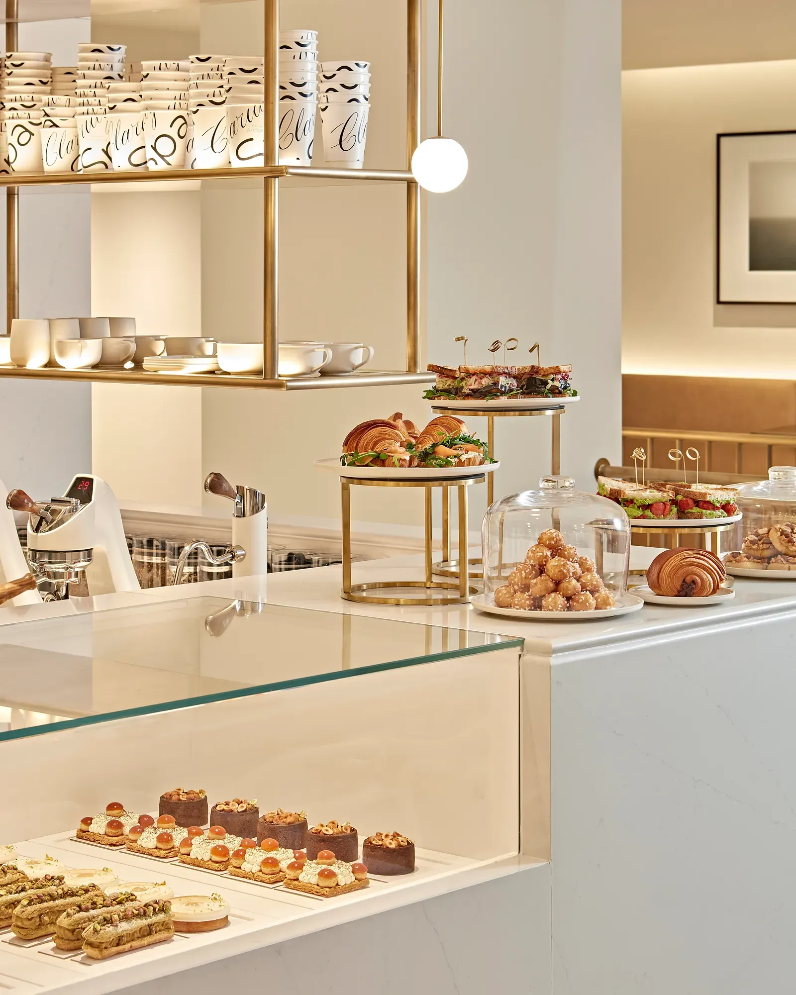 Bright and elegant patisserie counter displaying an assortment of pastries, cakes, and sandwiches on glass and tiered stands. The white and gold interior features modern shelving, ceramic cups, and soft lighting that highlights the refined presentation.