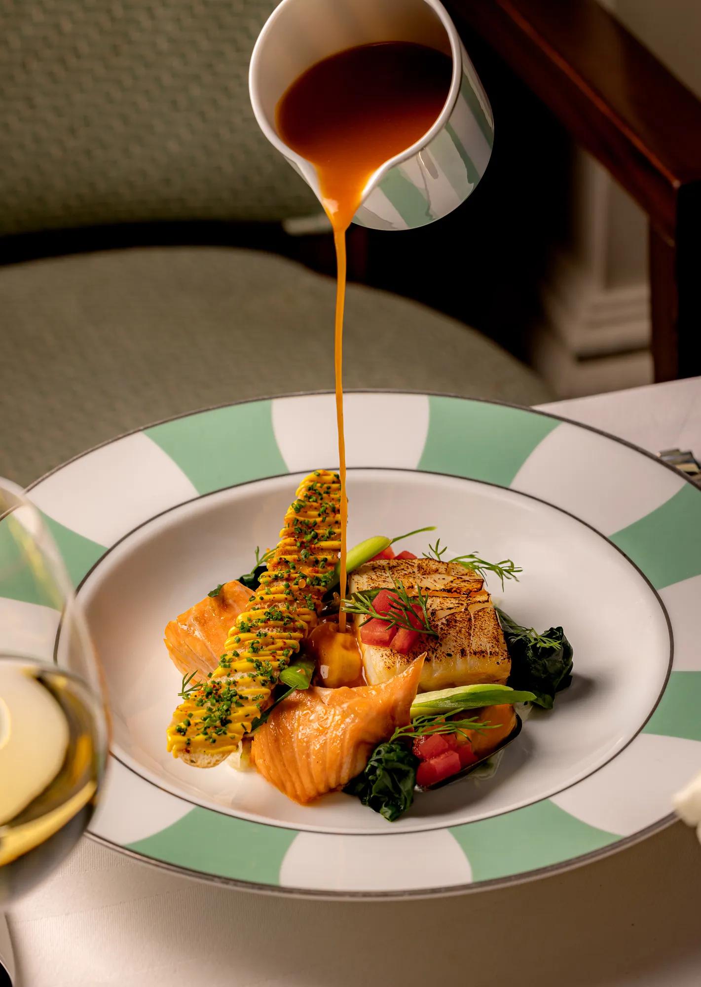 Sauce being poured over grilled salmon and vegetables on a green-striped plate.