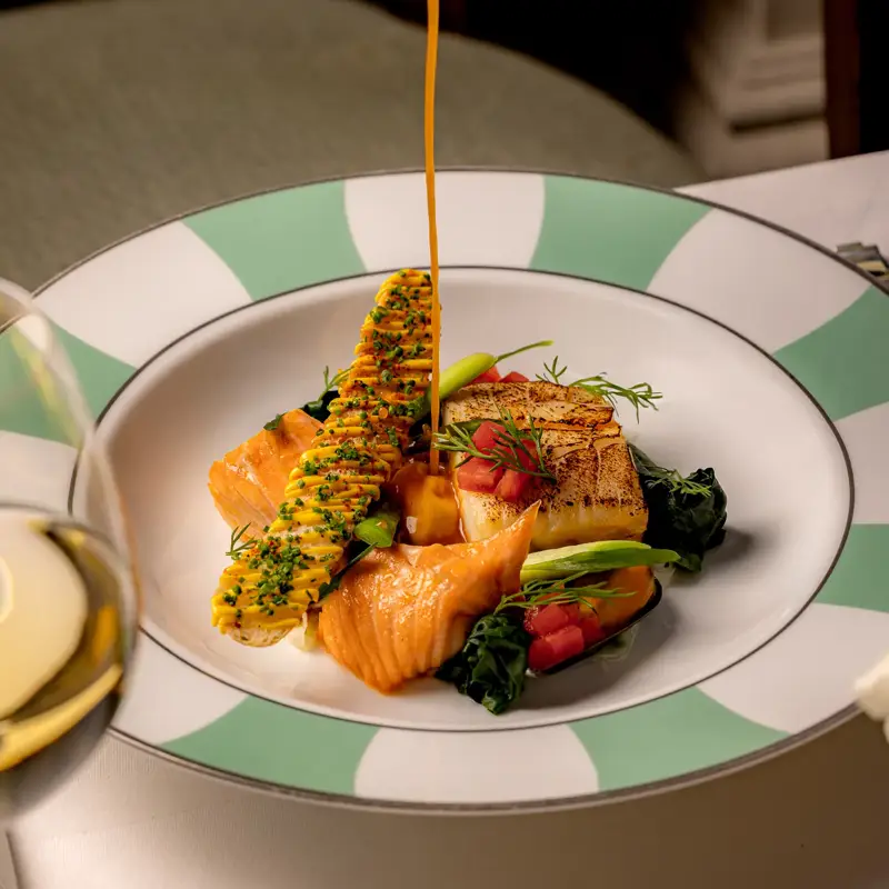 Sauce being poured over grilled salmon and vegetables on a green-striped plate.