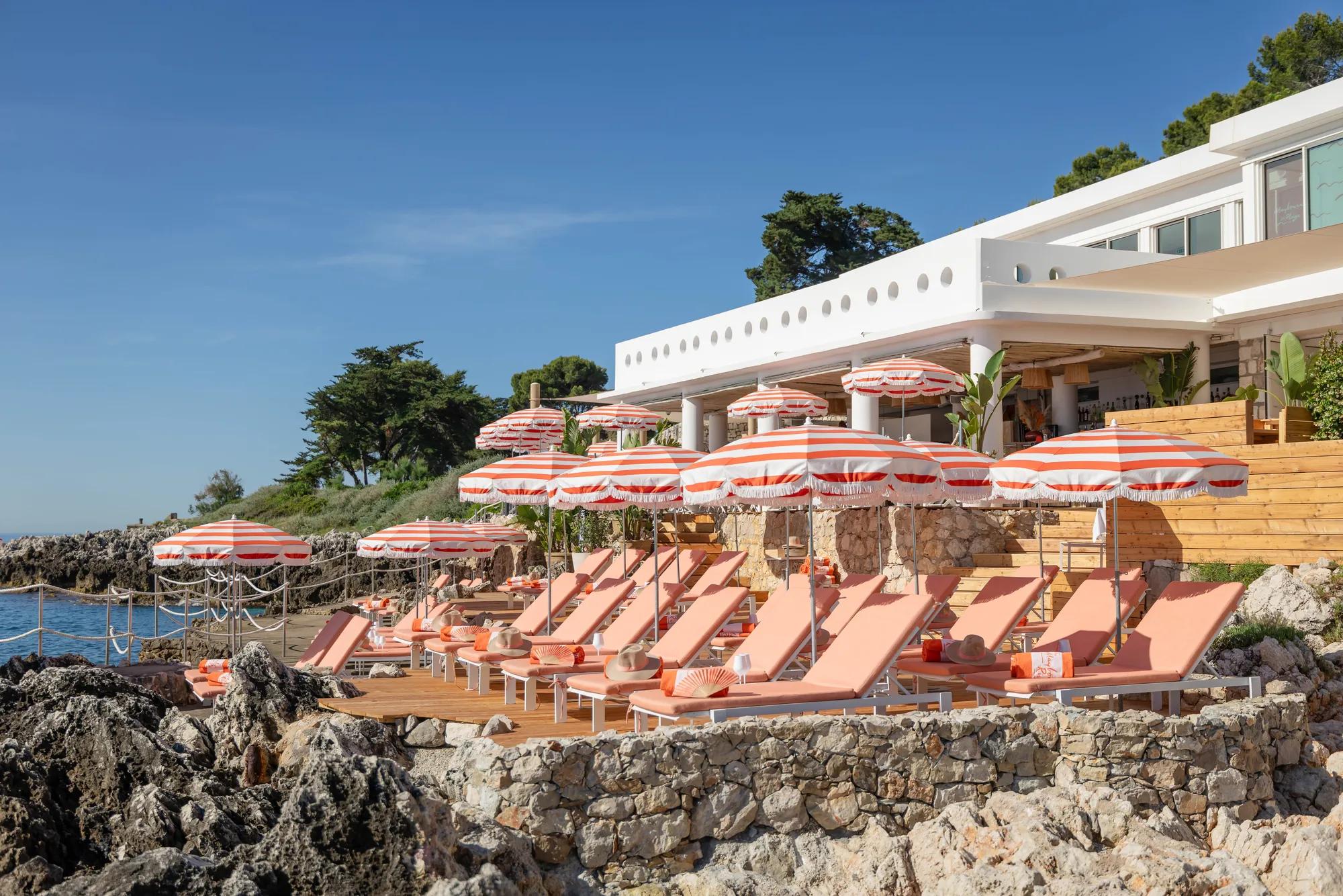 Transats et parasols rayés sur une terrasse en bord de mer, surplombant les rochers et la végétation côtière.