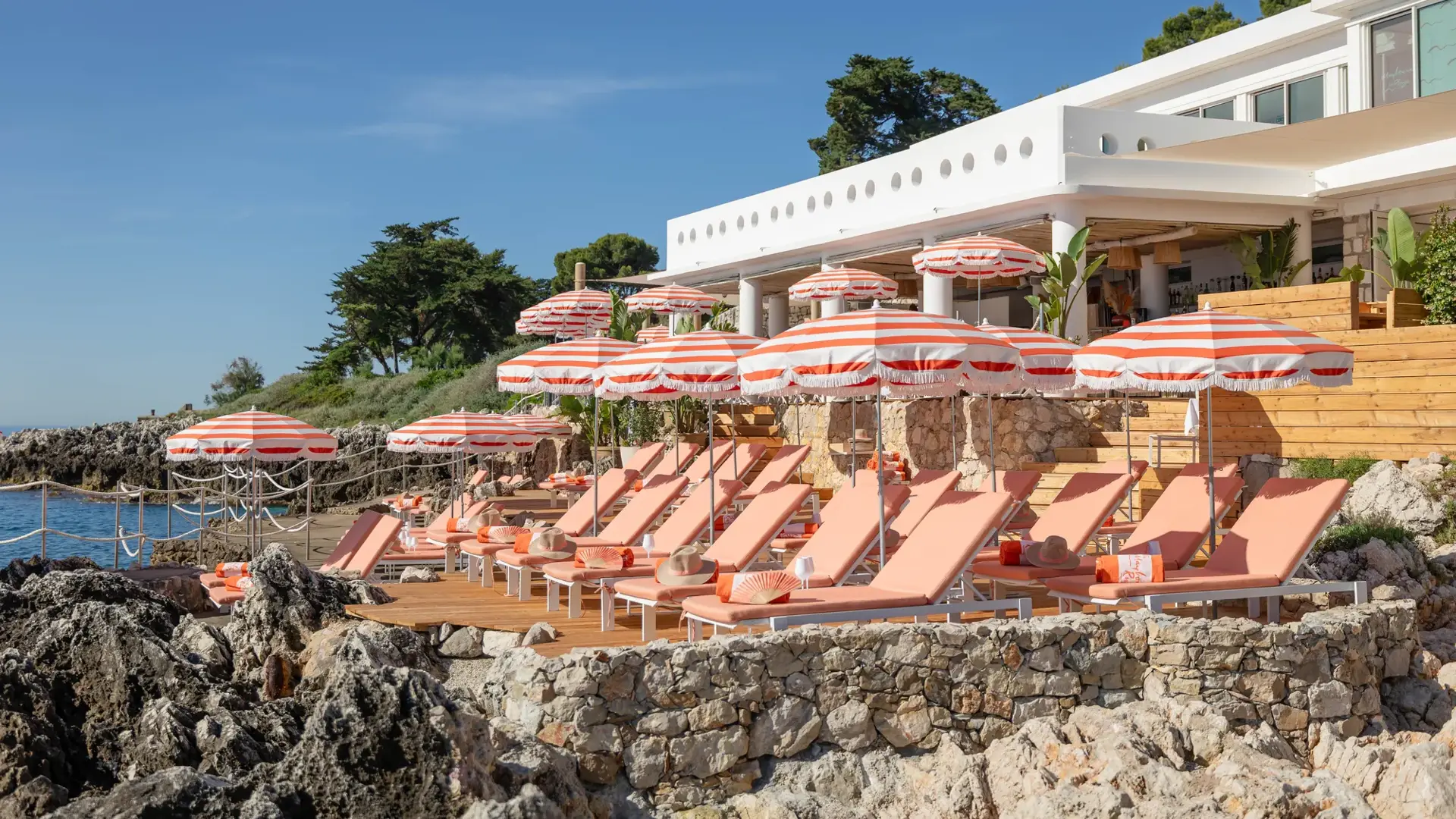 Sun loungers and striped umbrellas on a seaside terrace at a coastal restaurant with views of the rocky shore and trees.