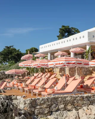 Transats et parasols rayés sur une terrasse en bord de mer, surplombant les rochers et la végétation côtière.