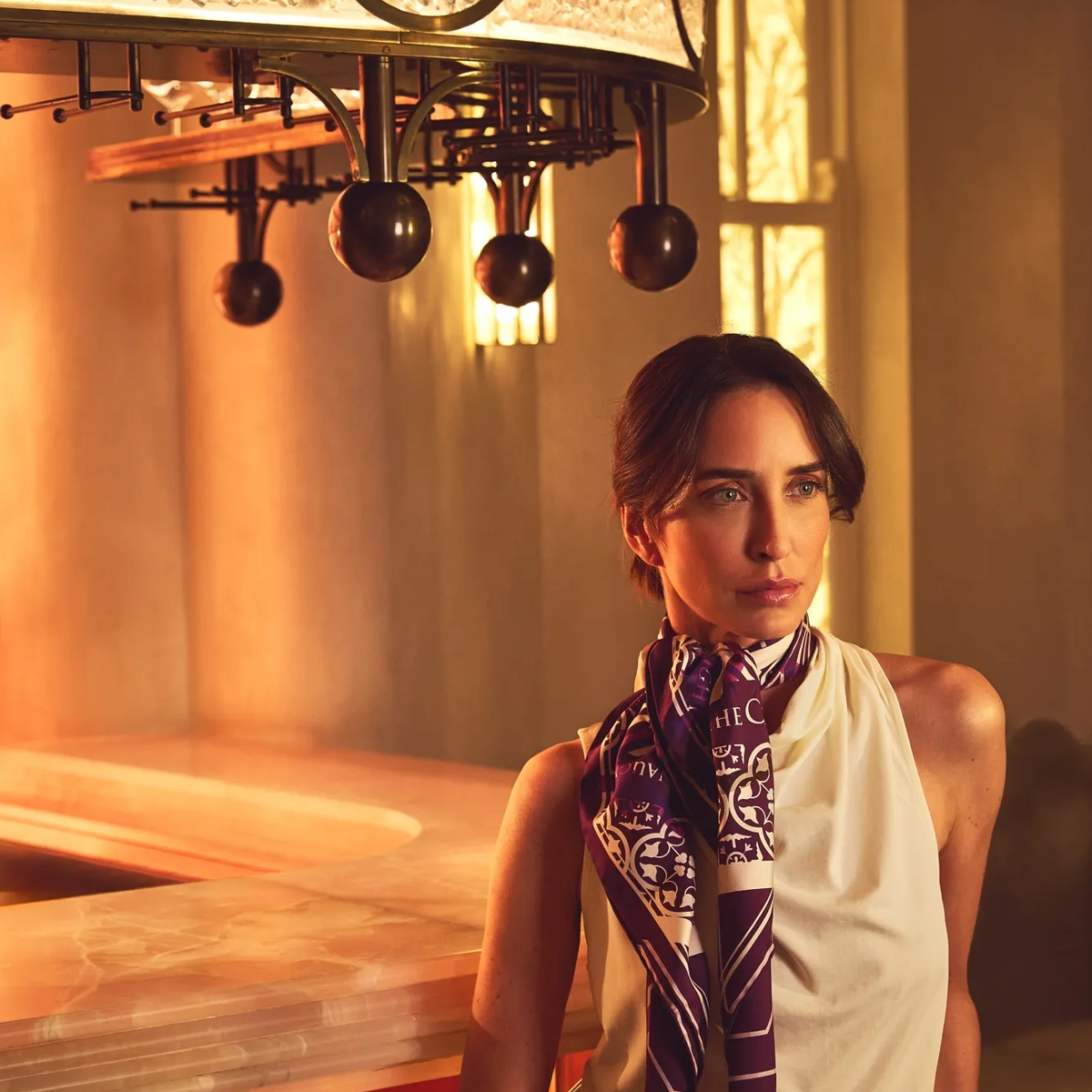 Guest seated at a softly lit bar wearing a silk scarf and sleeveless top, with warm marble surfaces and brass details behind.