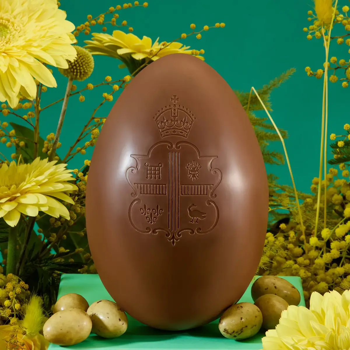 Large embossed chocolate Easter egg displayed on green pedestal surrounded by yellow flowers and small decorative eggs.
