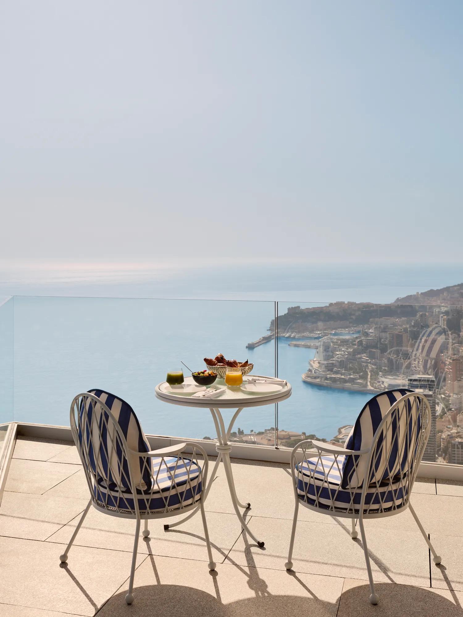 Elegant terrace setting with two blue-and-white chairs and a round table laid with drinks and snacks, overlooking the sea and Monaco’s coastline.