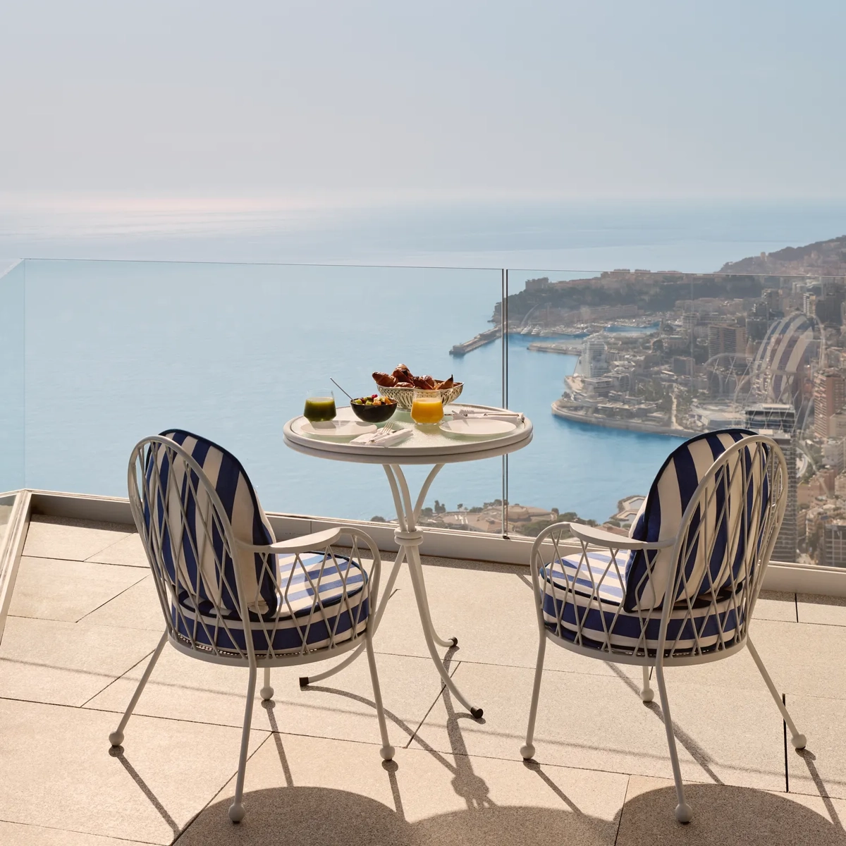 Elegant terrace setting with two blue-and-white chairs and a round table laid with drinks and snacks, overlooking the sea and Monaco’s coastline.