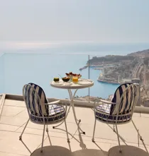 Elegant terrace setting with two blue-and-white chairs and a round table laid with drinks and snacks, overlooking the sea and Monaco’s coastline.