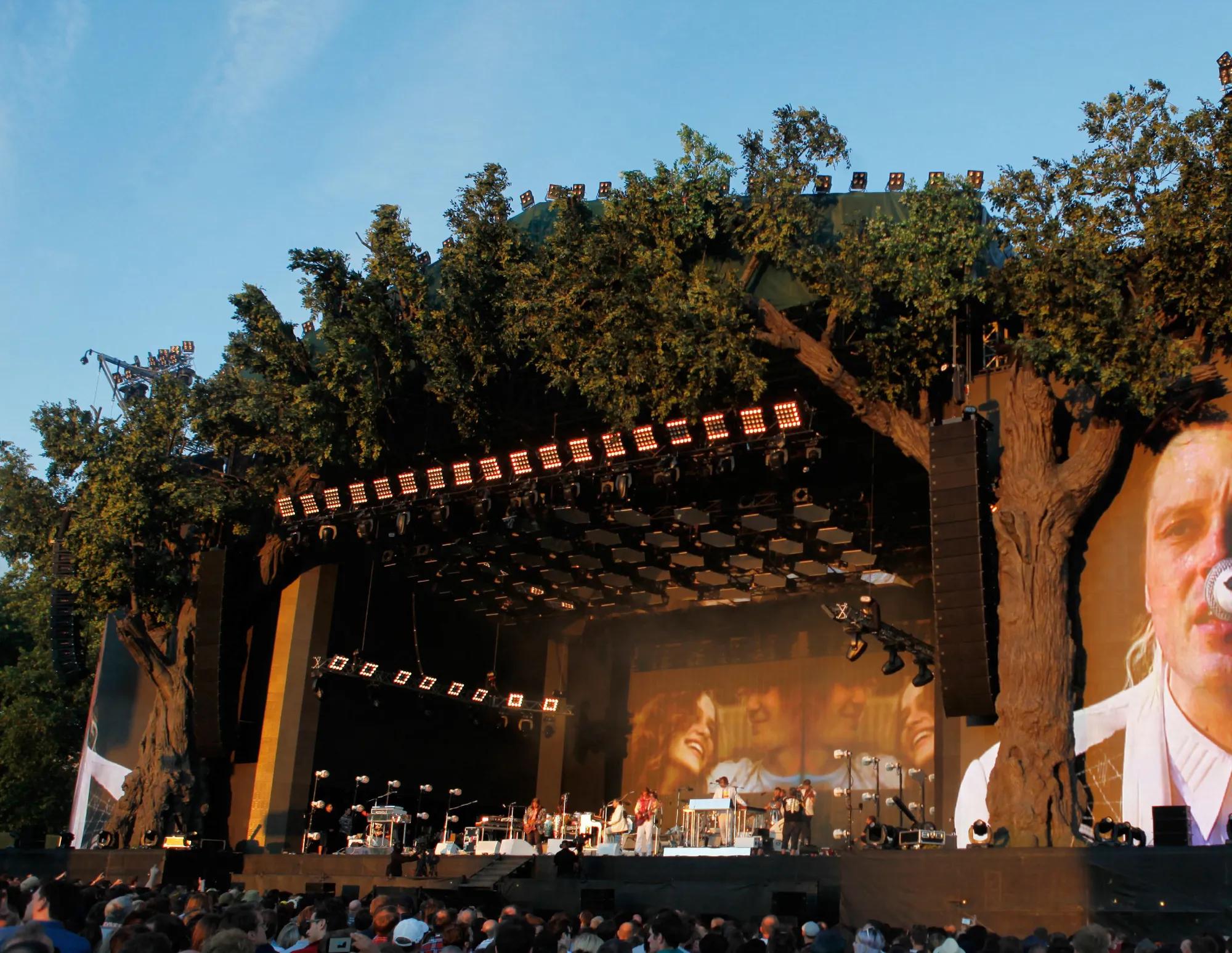 A photo of a big stage with musicians in a park with blue skies and a crowd of people watching