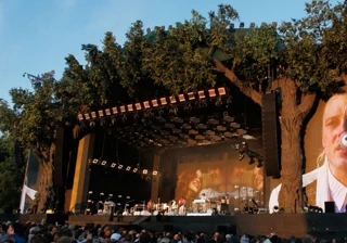 A photo of a big stage with musicians in a park with blue skies and a crowd of people watching