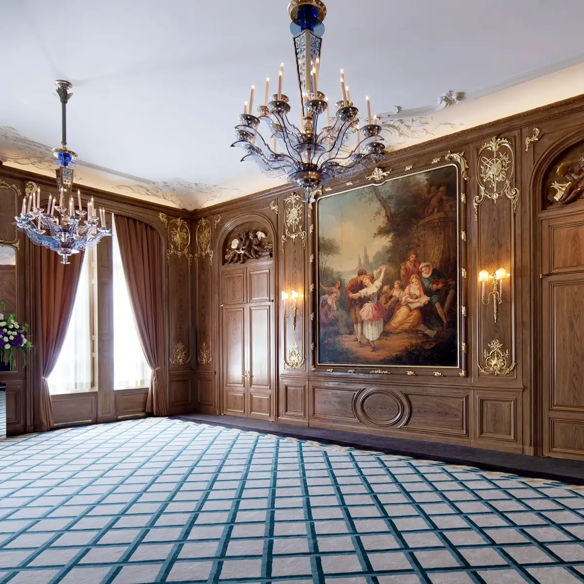 Elegant wood-panelled ballroom at Claridge’s featuring ornate chandeliers, a large classical painting framed in gold, draped windows, and a patterned blue and cream carpet.