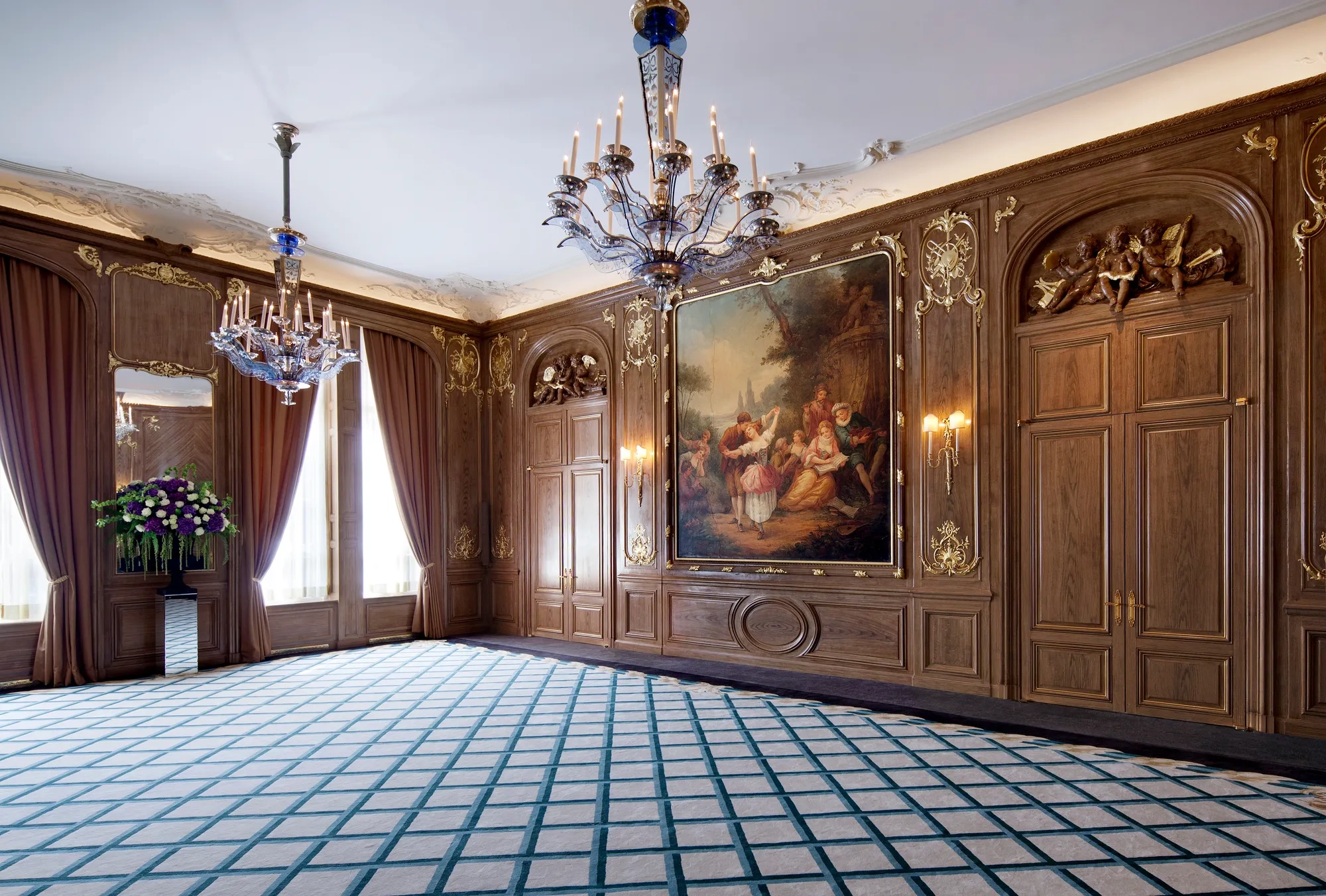 Elegant wood-panelled ballroom at Claridge’s featuring ornate chandeliers, a large classical painting framed in gold, draped windows, and a patterned blue and cream carpet.