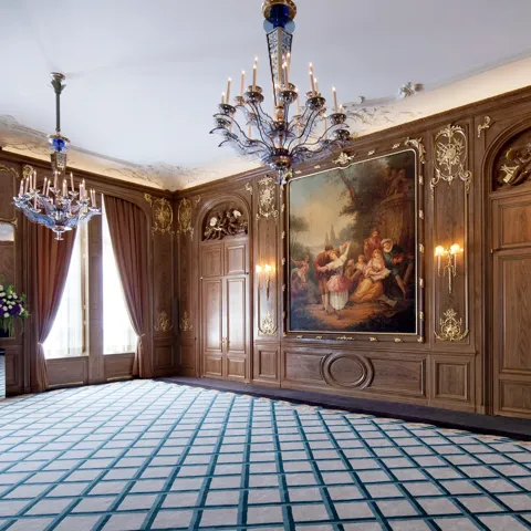 Elegant wood-panelled ballroom at Claridge’s featuring ornate chandeliers, a large classical painting framed in gold, draped windows, and a patterned blue and cream carpet.