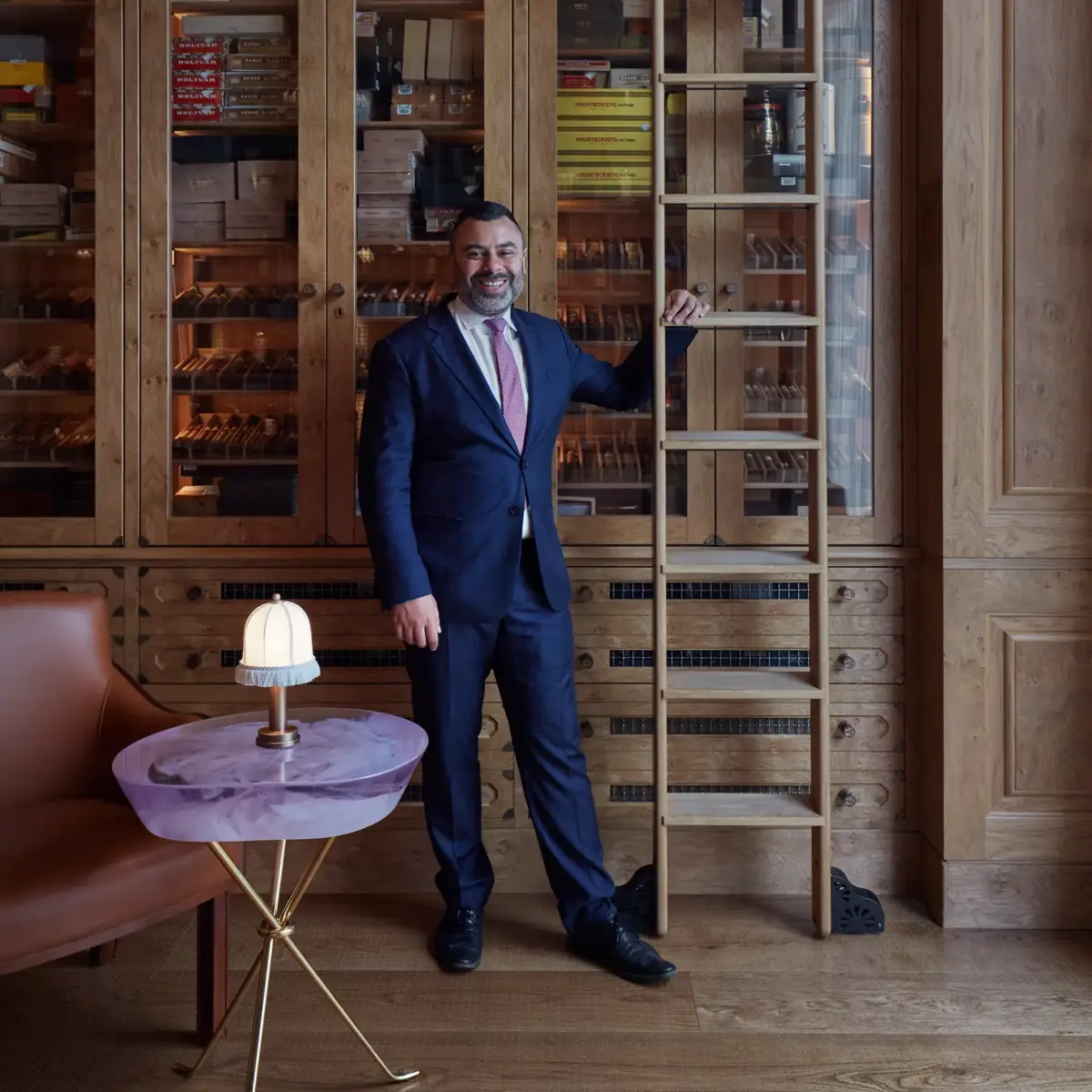 Man in a navy suit standing beside a wooden cigar cabinet and ladder in an elegant lounge.