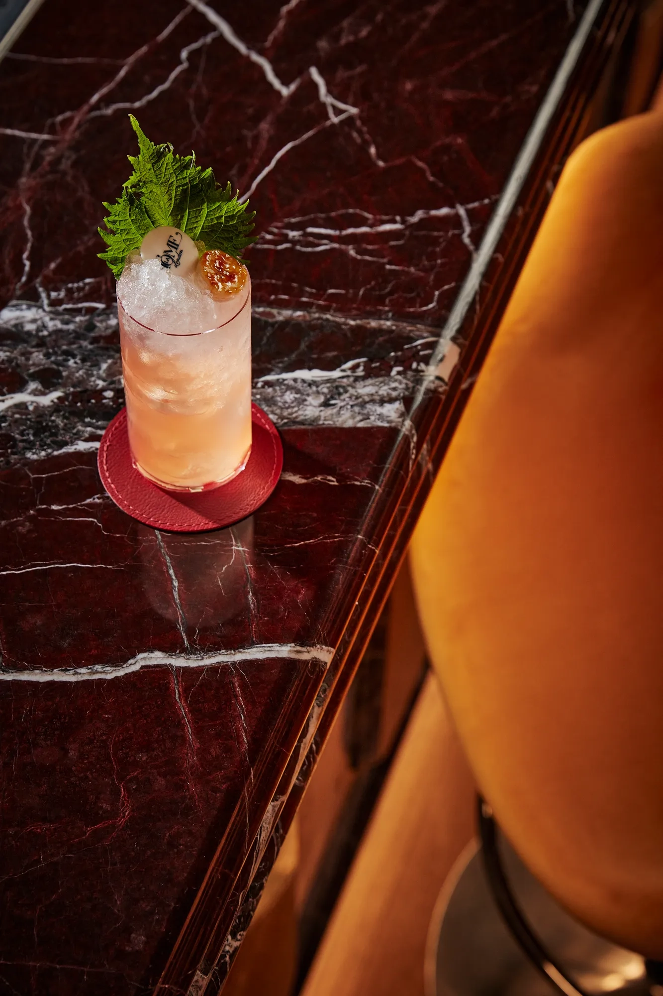 Tall cocktail with ice, garnish, and branded disc on dark red marble bar beside yellow chair.