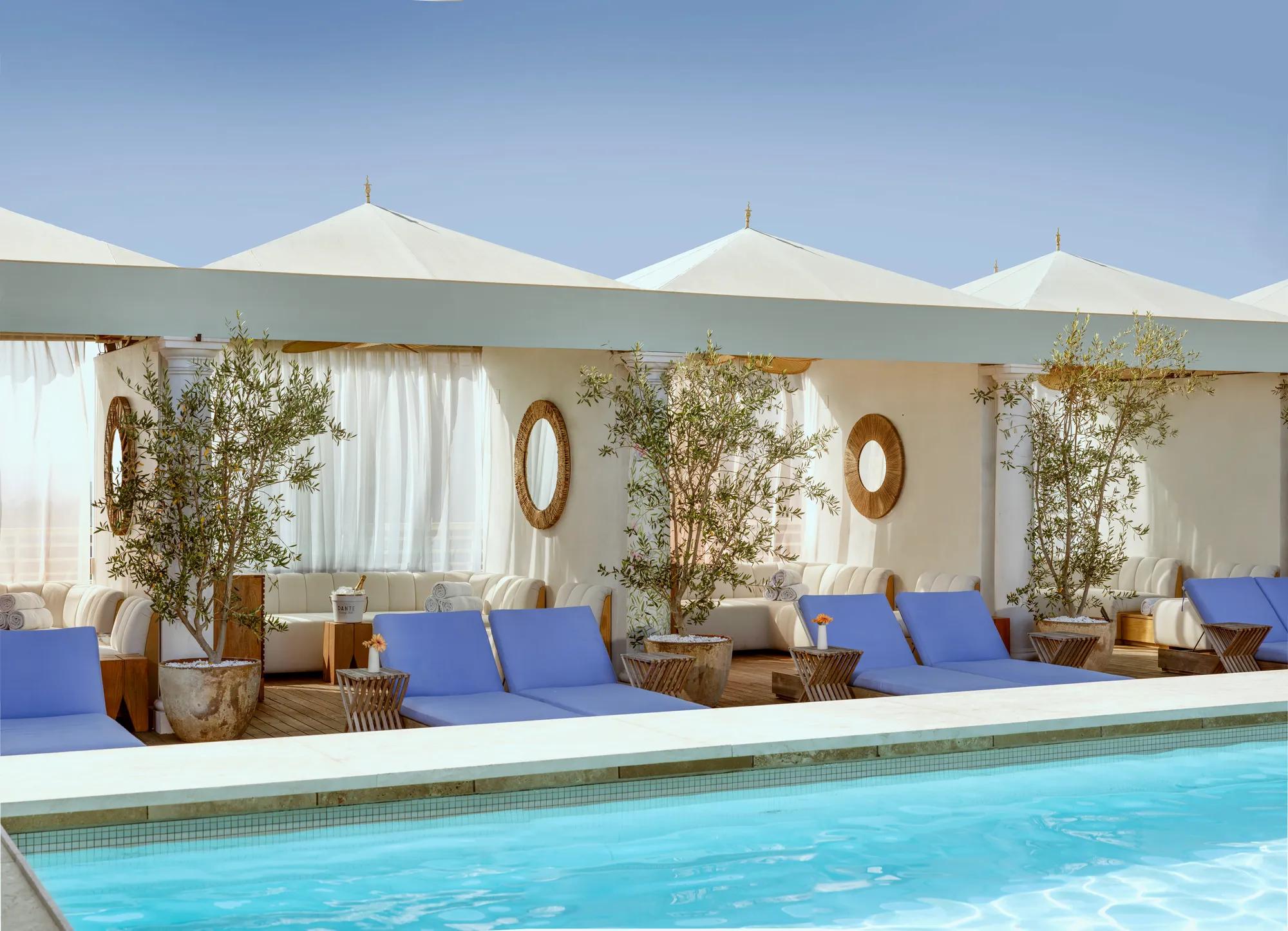Cabanas and blue loungers lining The Maybourne Beverly Hills rooftop pool beneath white canopies.