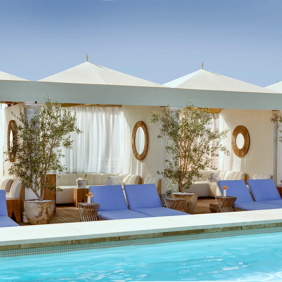 Cabanas and blue loungers lining The Maybourne Beverly Hills rooftop pool beneath white canopies.