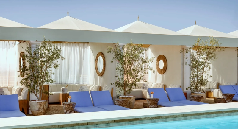 Cabanas and blue loungers lining The Maybourne Beverly Hills rooftop pool beneath white canopies.
