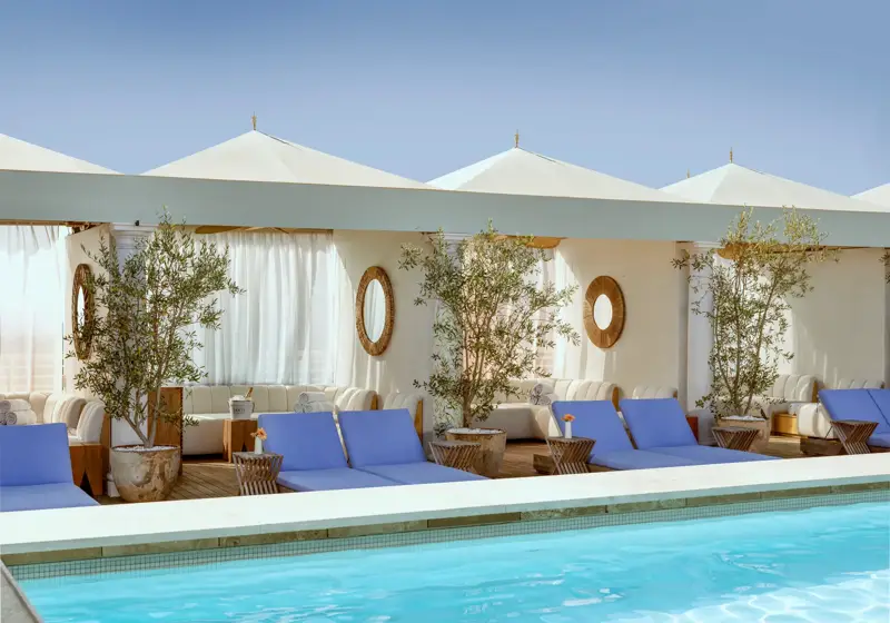 Cabanas and blue loungers lining The Maybourne Beverly Hills rooftop pool beneath white canopies.