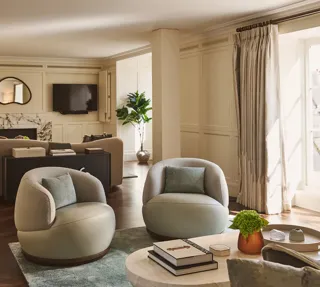 Elegant lounge with soft cream walls, round armchairs, marble fireplace, and large windows letting in natural light.