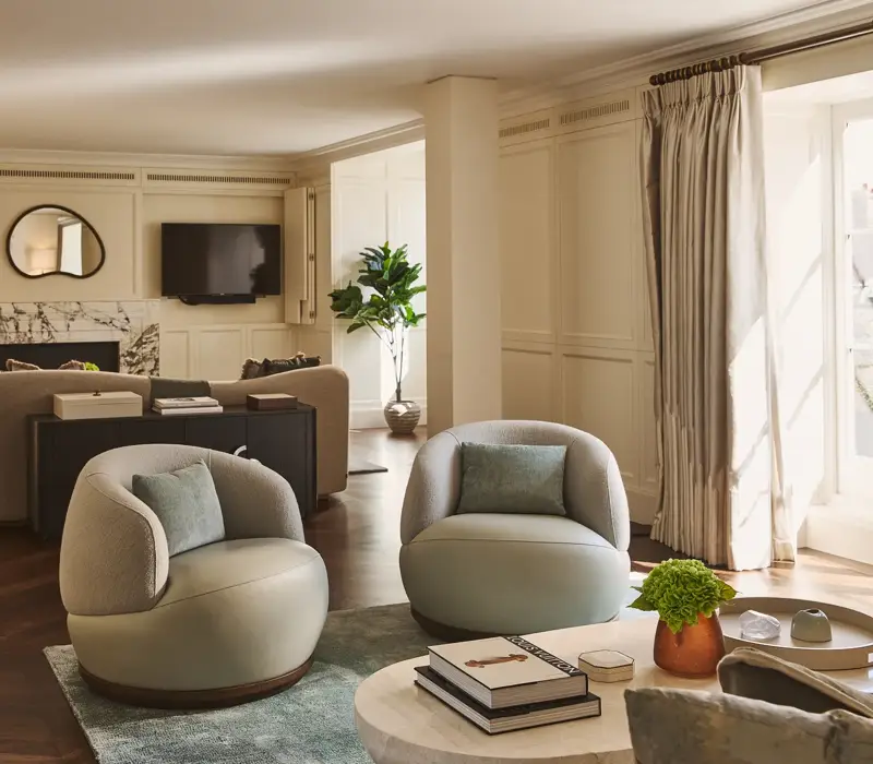 Elegant lounge with soft cream walls, round armchairs, marble fireplace, and large windows letting in natural light.