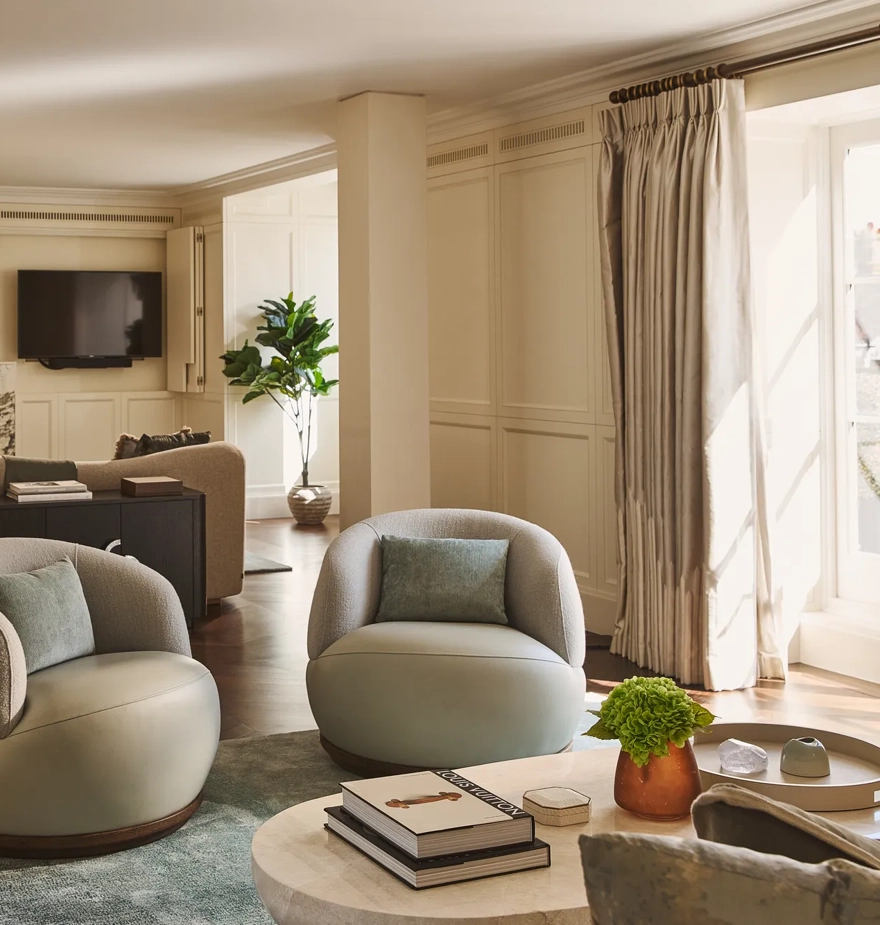 Elegant lounge with soft cream walls, round armchairs, marble fireplace, and large windows letting in natural light.