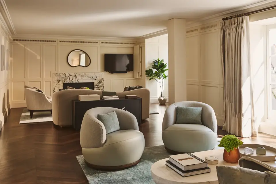 Elegant lounge with soft cream walls, round armchairs, marble fireplace, and large windows letting in natural light.
