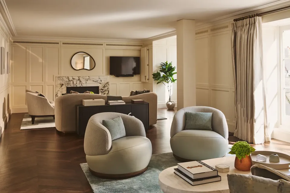 Elegant lounge with soft cream walls, round armchairs, marble fireplace, and large windows letting in natural light.