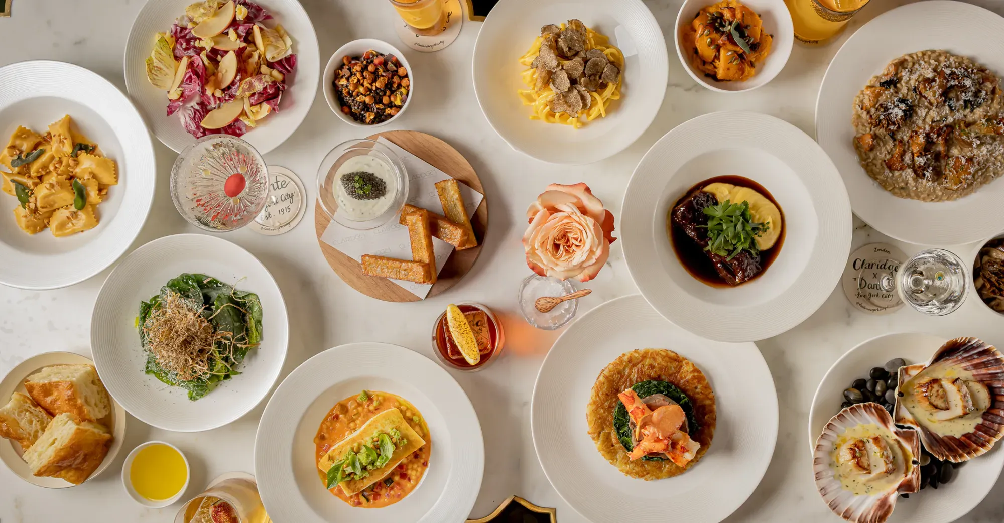 Overhead view of a table set with plated dishes, pasta, seafood, salads, bread, and drinks arranged for shared dining.