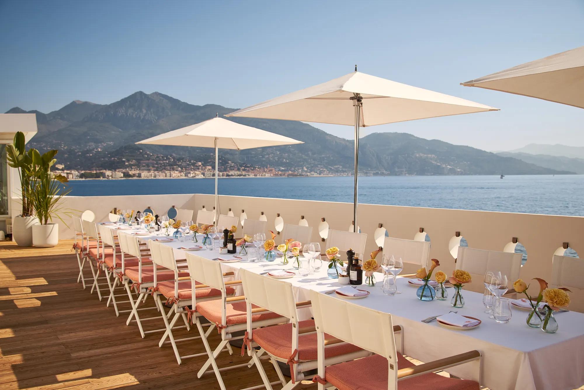 Longue table dressée en terrasse avec parasols blancs, fleurs et verrerie, donnant sur la mer et les montagne