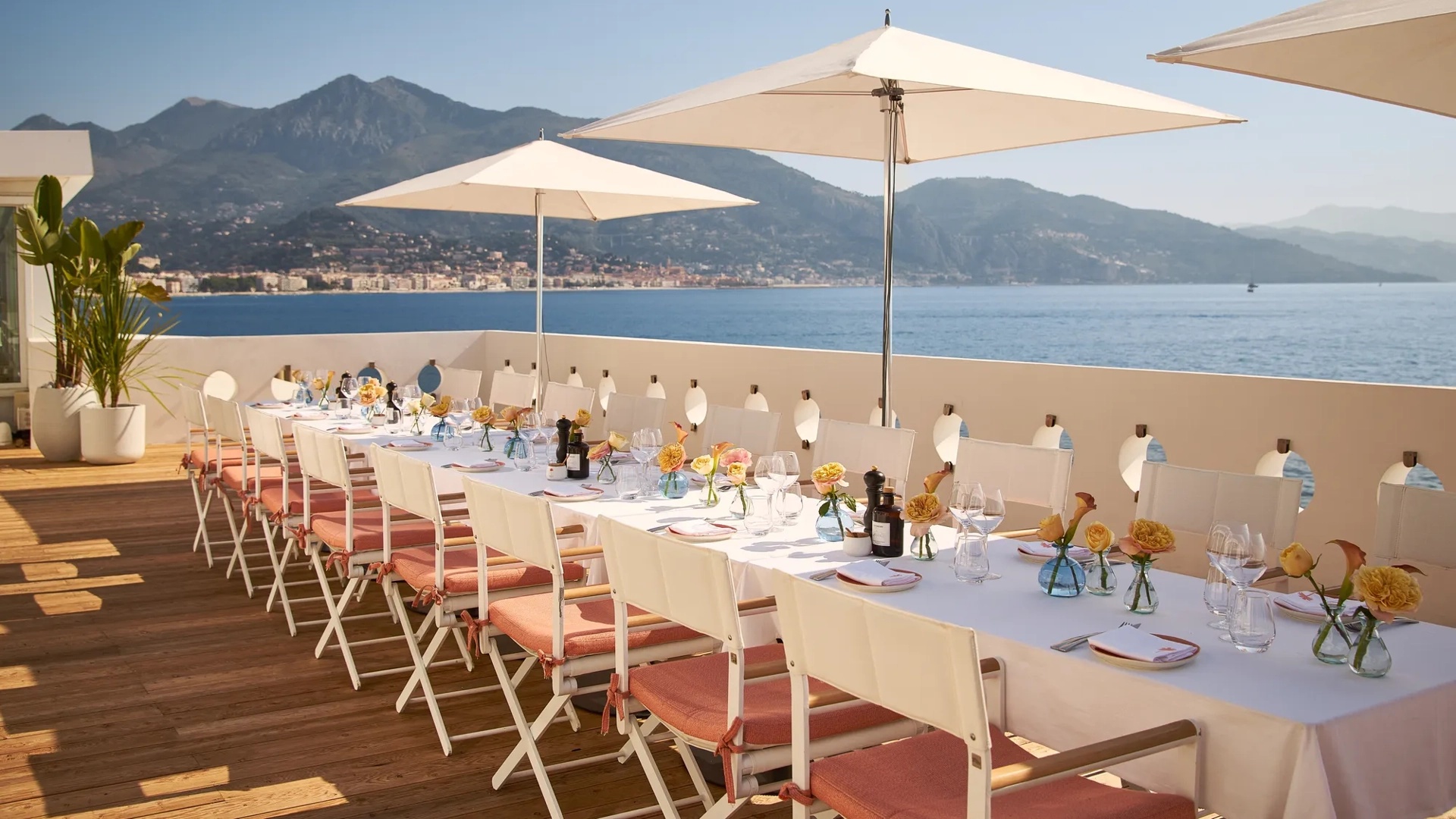 Long terrace dining table with white umbrellas, set with flowers and glassware overlooking sea and mountains