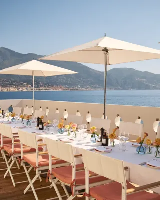 Longue table dressée en terrasse avec parasols blancs, fleurs et verrerie, donnant sur la mer et les montagne