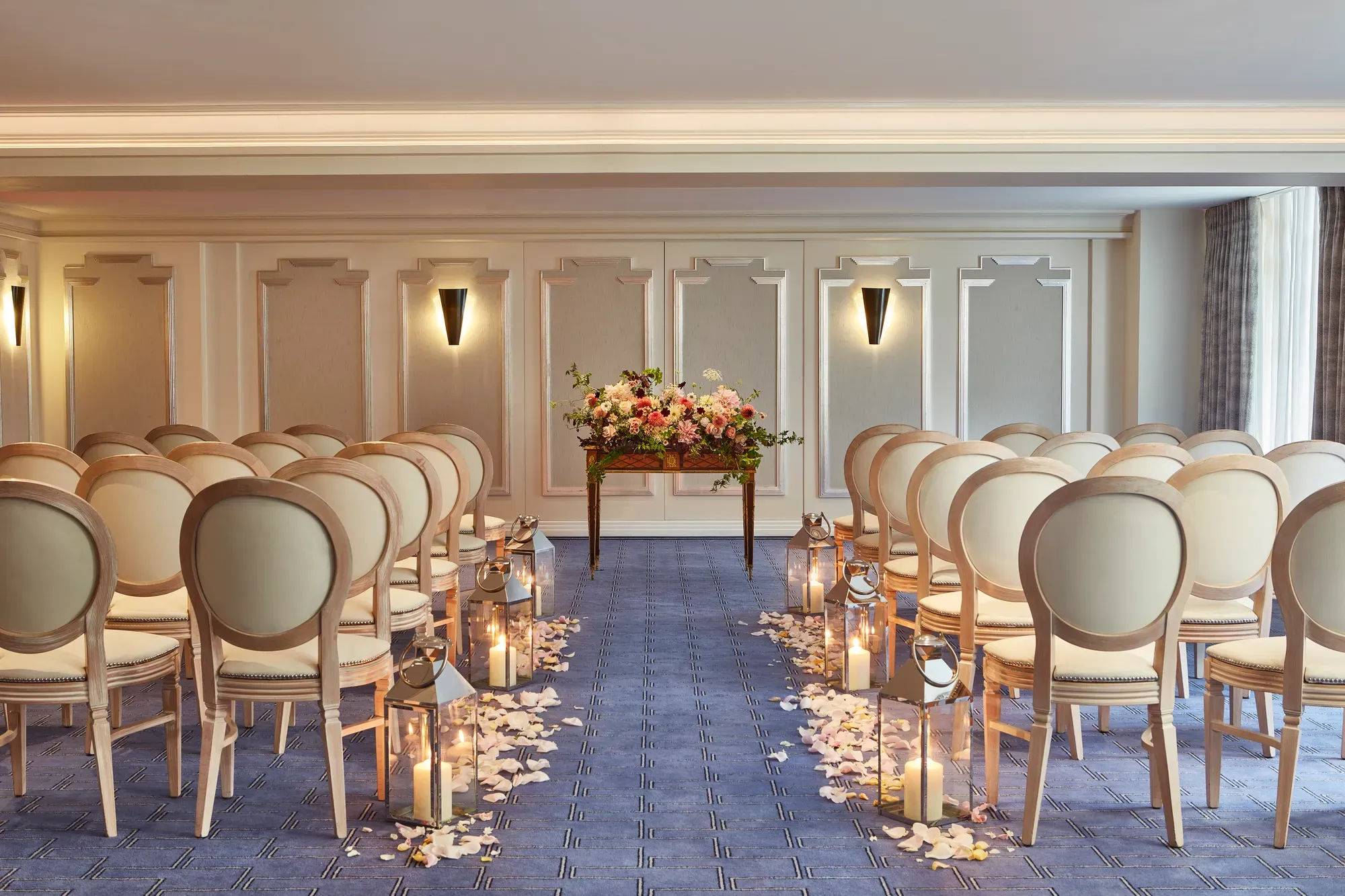 Elegant wedding ceremony setup at Claridge’s with cream chairs arranged in rows, an aisle lined with candles and flower petals, and a floral arrangement displayed on a wooden table at the front.