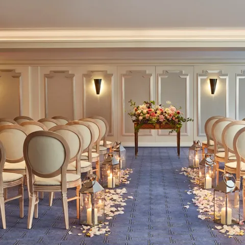 Elegant wedding ceremony setup at Claridge’s with cream chairs arranged in rows, an aisle lined with candles and flower petals, and a floral arrangement displayed on a wooden table at the front.